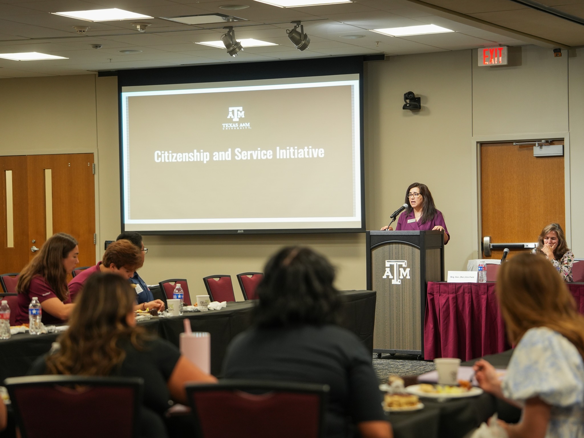 Dr. Gonzalez at a podium presents to a seated audience, with a screen displaying "Citizenship and Service Initiative" at Texas A&M University.